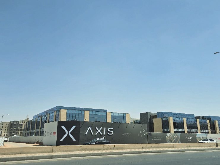Glass-and-stone office blocks of Axis Commercial Center rising behind branded site hoarding